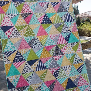 May include: A colorful quilt with a geometric pattern of triangles in shades of blue, green, pink, yellow, and white. The quilt is made of various fabrics with different patterns, including polka dots, stripes, and floral designs. The quilt is hanging on a rope and the text "Stereo" is visible at the top of the image. The text "Jaybird Quilts" is visible at the bottom of the image.