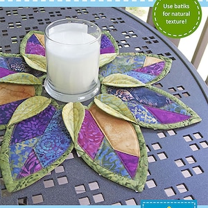 May include: A leaf-shaped table topper with a candle in the center. The topper is made of colorful batik fabric in shades of purple, blue, and gold, with green edges. The text "FOLD'N STITCH LEAF TOPPER" is at the top. The finished size is 17 inches in diameter.