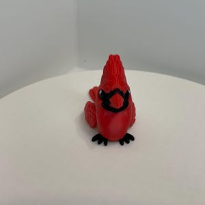 May include: A red cardinal figurine with black accents. The bird is perched on a white surface. The cardinal has a detailed crest, black mask, and black feet. The figurine is a decorative item.
