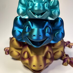 May include: Three stacked 3D printed figurines of a smiling cartoon creature. The top figurine is teal blue, the middle figurine is blue, and the bottom figurine is gold with a hint of purple.