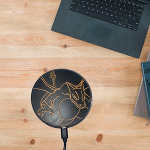 May include: A black wireless charging pad with a gold outline of a dragon design. The charging pad is on a wooden surface.
