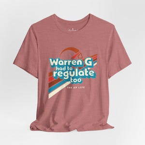 May include: A dusty rose t-shirt with the text "Warren G had to regulate too" in a retro font. The design includes a sun and colorful stripes. The text "THE AP LIFE" is below the main text.