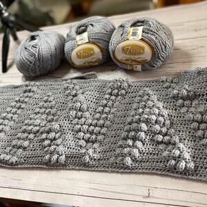 May include: A gray crocheted blanket with a textured pattern. Three balls of gray yarn are in the background, one with the label "Trego" on it.