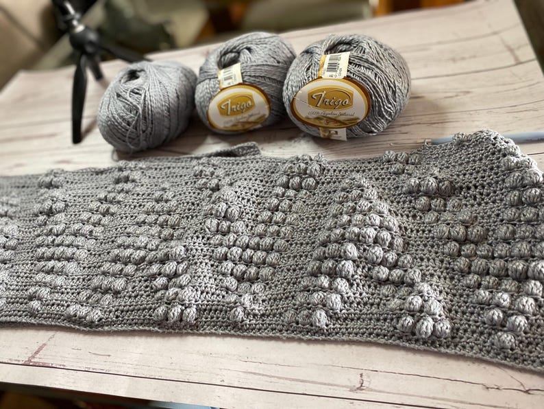 May include: A gray crocheted blanket with a textured pattern. Three balls of gray yarn are in the background, one with the label "Trego" on it.