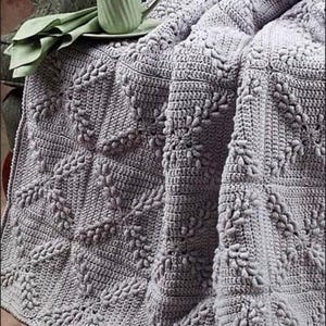 May include: A soft, gray crocheted throw blanket with a textured, floral pattern. The blanket is draped over a chair and a green teacup is sitting on a table in the background.