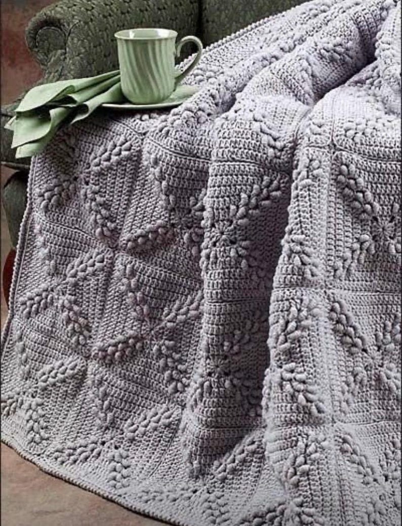 May include: A soft, gray crocheted throw blanket with a textured, floral pattern. The blanket is draped over a chair and a green teacup is sitting on a table in the background.