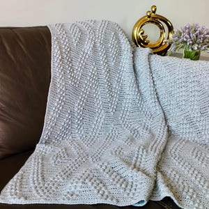 May include: A soft, gray crocheted throw blanket with a textured, raised pattern. The blanket is draped over a brown leather couch.