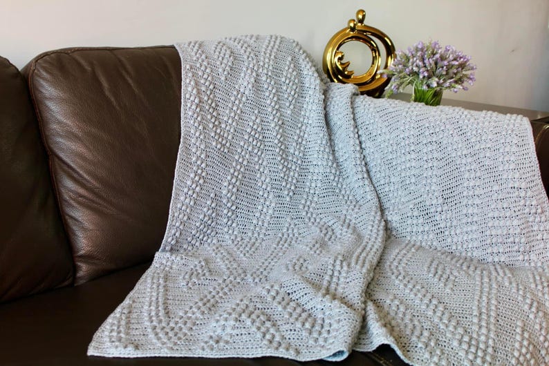 May include: A soft, gray crocheted throw blanket with a textured, raised pattern. The blanket is draped over a brown leather couch.
