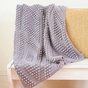 May include: A soft, gray crocheted throw blanket with a textured, bobble stitch pattern. The blanket is draped over a white bench with a yellow pillow.