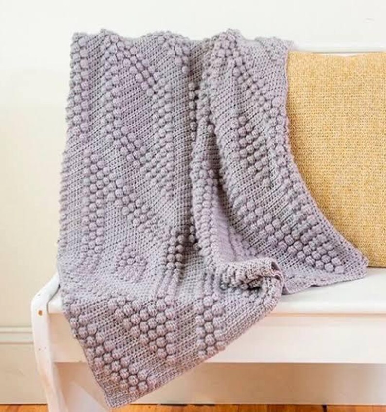May include: A soft, gray crocheted throw blanket with a textured, bobble stitch pattern. The blanket is draped over a white bench with a yellow pillow.