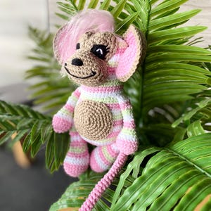May include: A handmade crochet monkey doll with a pink and white striped body, pink ears, and a pink hair tuft. The monkey has a tan face and a long pink tail. The doll is surrounded by green leaves.
