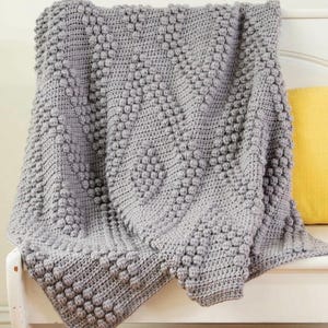 May include: A gray crocheted throw blanket with a textured bobble stitch pattern. The blanket is draped over a white bench.