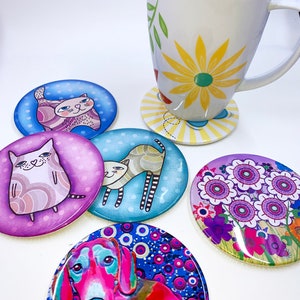 Puede incluir: Una taza blanca con un dise&ntilde;o floral amarillo se encuentra sobre un posavasos a juego. Varios posavasos redondos y coloridos con ilustraciones de gatos y perros est&aacute;n dispersos. Los posavasos presentan colores vibrantes y dise&ntilde;os &uacute;nicos.