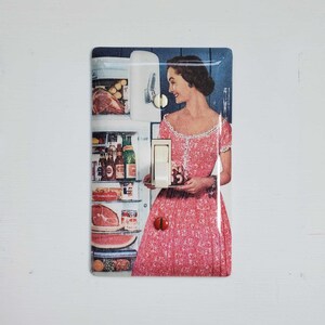 May include: A vintage-style light switch cover featuring a woman in a pink dress standing in front of a refrigerator filled with groceries. The switch cover has a white toggle switch and is made of metal.