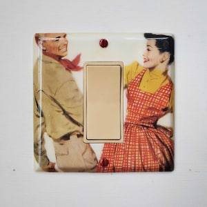 May include: A light switch cover featuring a vintage image of a couple dancing. The woman is wearing a red and white gingham dress and the man is wearing a brown jacket and khaki trousers.