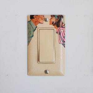 May include: A light switch cover featuring a retro illustration of a couple on the verge of a kiss. The artwork depicts a man in a green shirt and a woman in a striped pink and white top. The switch and screws are silver.