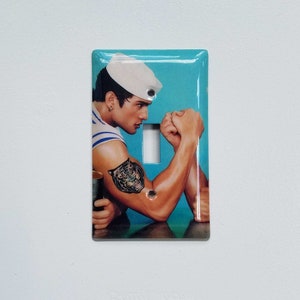 May include: A light blue light switch cover featuring a photograph of a man wearing a sailor hat and a tiger tattoo on his arm. The man is flexing his arm, seemingly engaged in an arm wrestle.