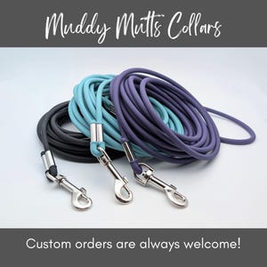 May include: Three dog leashes in shades of gray, teal, and purple, coiled and ready for use. Each leash has a silver metal clasp. The text "Muddy Mutts Collars" is at the top, and "Custom orders are always welcome!" is at the bottom.