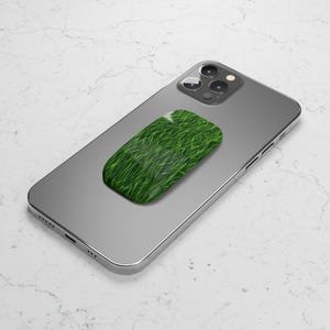 May include: A smartphone with a green grass-textured phone grip attached to the back. The grip is rectangular and features a vibrant green grass design. The phone is silver and has a triple-lens camera.