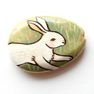 May include: An oval-shaped stone painted with a white rabbit running through a light green grassy field. The rabbit has long ears, pink cheeks, and dark outlines. The background features subtle green grass blades and small pink dots.