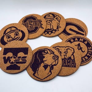 May include: Nine round cork coasters with various college logos and mascots, including Ohio State, Georgia, Alabama, and Tennessee. The coasters are all brown and have black designs.