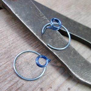 May include: Two blue and silver hoop earrings with a spiral design. The earrings are set against a background of a metal tool and a wooden surface. The earrings are a simple, yet elegant design.