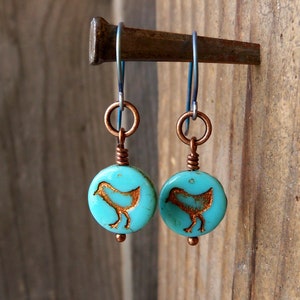 May include: A pair of turquoise blue earrings with copper wire and a copper bird design. The earrings are hanging from a silver hook.