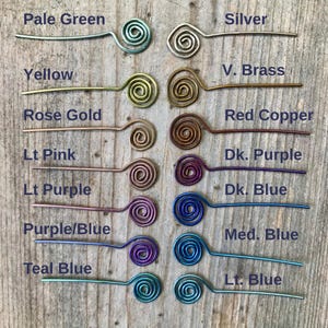 May include: A collection of wire hair pins in various colors, including pale green, silver, yellow, rose gold, and red copper. Each pin features a spiral design at one end and is displayed on a wooden surface.