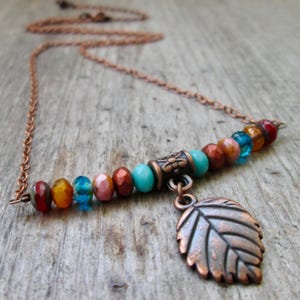 May include: A copper-toned necklace featuring a bar of colorful beads in shades of red, orange, blue, and brown. A copper leaf charm hangs from the bar. The necklace chain is also copper-toned.