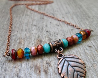 May include: A copper-toned necklace featuring a bar of colorful beads in shades of red, orange, blue, and brown. A copper leaf charm hangs from the bar. The necklace chain is also copper-toned.