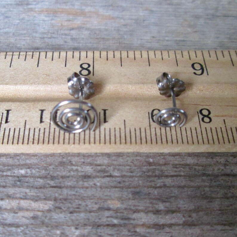 May include: A pair of silver-toned spiral stud earrings. The earrings have a post and butterfly backing. The spiral design is a simple, elegant shape. The earrings are shown on a wooden surface with a ruler.