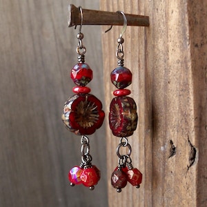 May include: Red and gold glass bead earrings with a floral design. The earrings are hanging from silver hooks and have three smaller red beads dangling from the bottom.
