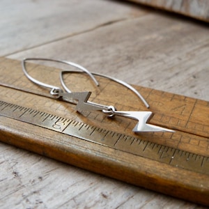 May include: A pair of silver lightning bolt earrings with a simple, minimalist design. The earrings are hanging from a wooden ruler.
