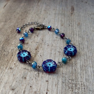 May include: A blue and purple beaded bracelet with a silver chain and three flower-shaped charms. The charms have a blue and white floral design.