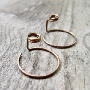 May include: Two copper-colored hoop earrings with a unique design. Each earring features a circular hoop connected to a small, coiled accent. The earrings are displayed on a weathered, gray wooden surface.