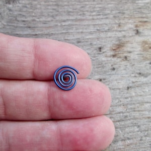 May include: A small, spiral-shaped, blue metal object. The spiral is tightly wound, with a slightly open end. The object is resting on a person's fingers, with a wooden surface in the background.