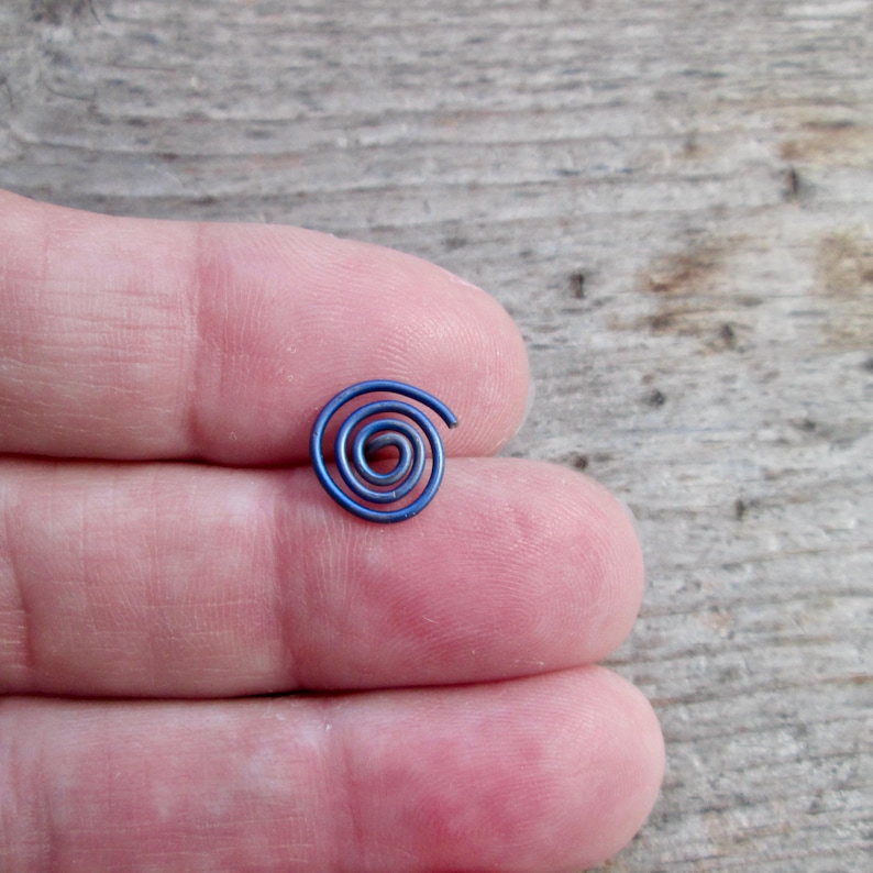 May include: A small, spiral-shaped, blue metal object. The spiral is tightly wound, with a slightly open end. The object is resting on a person's fingers, with a wooden surface in the background.