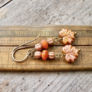 May include: A pair of dangle earrings with peach and orange glass beads and peach leaf charms. The earrings are hanging on a wooden ruler.