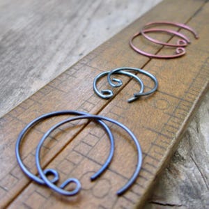 May include: Three pairs of handcrafted hoop earrings in shades of blue, gray, and pink. The earrings are made of thin metal wire and have a unique, open-ended design with a looped detail. The earrings are displayed on a wooden surface.