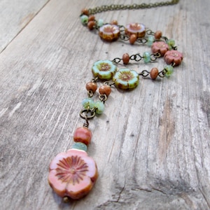 May include: A floral necklace with a vintage aesthetic. The necklace features pink, blue, and brown flower-shaped beads, interspersed with smaller beads in complementary colors. The chain is a dark bronze color, and the necklace is displayed on a wooden surface.