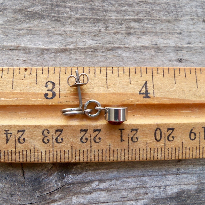 May include: A pair of silver earrings with a red gemstone dangle. The earrings are measured against a wooden ruler, showing they are just over 3 inches long.