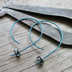 May include: A pair of teal-colored hoop earrings. The earrings are made of thin, curved metal and have a post and backing. The background is a wooden surface with a metal tool.