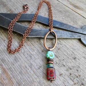 May include: A copper-toned necklace with an oval pendant and a chain. The pendant features a turquoise bead, brown and blue beads, and a red rectangular bead. The necklace is displayed on a wooden surface.