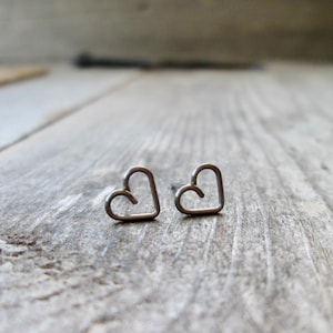 May include: A pair of small, silver-colored heart-shaped stud earrings. The earrings are simple in design, with a minimalist, open-heart outline. The earrings are displayed on a weathered, gray wooden surface.