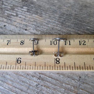 May include: Two small, silver-colored stud earrings with a circular design. The earrings are shown on a wooden surface next to a ruler, with the earring posts and backs visible. The earrings are approximately 0.4 inches in diameter.