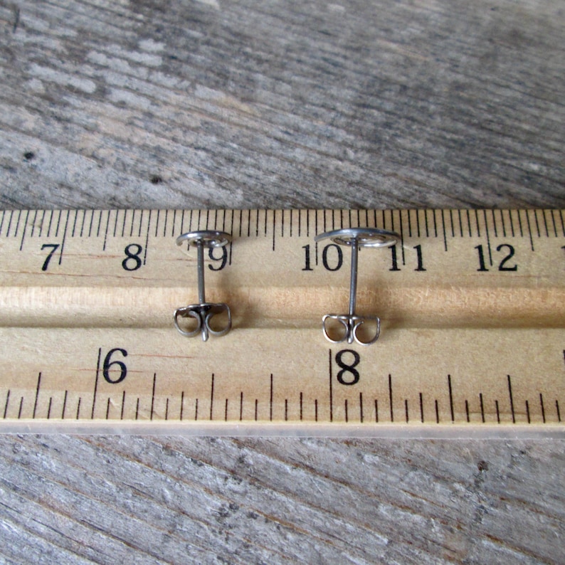 May include: Two small, silver-colored stud earrings with a circular design. The earrings are shown on a wooden surface next to a ruler, with the earring posts and backs visible. The earrings are approximately 0.4 inches in diameter.