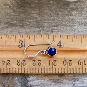 May include: A close-up of a blue crystal earring with a silver hook. The earring is about 1/2 inch long and is shown next to a ruler.