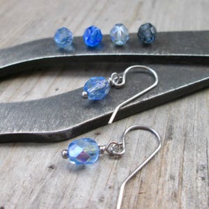May include: A pair of silver earrings with blue iridescent glass beads. The earrings are on a metal surface.
