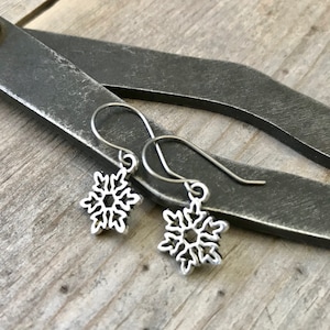 May include: A pair of silver snowflake earrings with delicate wire loops. The earrings are small and dainty, perfect for adding a touch of winter charm to any outfit.