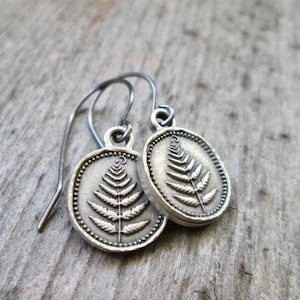 May include: A pair of silver-toned drop earrings featuring a circular pendant with a raised fern leaf design. The edges of the circular charms are detailed with a beaded texture, and the earrings hang from simple curved silver-toned metal hooks.